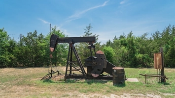 Idle pumpjack on dry grass with a rusted barrel at its base, set against a backdrop of dense green trees